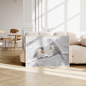 May include: A framed painting depicting two birds perched on a snowy branch. The birds are grey and white with brown accents. The background is a soft grey with a subtle snow effect.