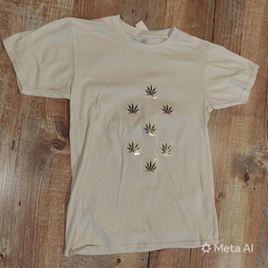 May include: A white short-sleeved t-shirt featuring a central graphic pattern composed of six individual dark green cannabis leaf icons arranged in a diamond-like formation on the front chest area of the garment.