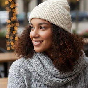 Puede incluir: Una mujer con un gorro de punto acanalado color crema y una bufanda y suéter de punto gris. El texto "Yelé Silk Lined Beanies" está en la parte inferior de la imagen. El fondo está borroso con luces bokeh.