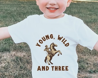 Könnte beinhalten: Ein kleiner Junge trägt ein weißes T-Shirt mit einem braunen Aufdruck eines Cowboys, der ein Pferd reitet. Das Shirt sagt "Young, Wild and Three".