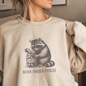 Peut inclure: Un sweat-shirt beige avec un imprimé d'un raton laveur tenant un pot de cornichons. Le texte "Never Enough Pickles" est imprimé sous l'imprimé.