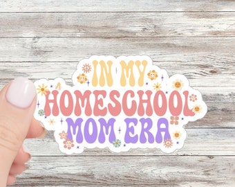 Puede incluir: Una pegatina blanca con el texto "In My Homeschool Mom Era" en una fuente retro. El texto está en amarillo, rosa y morado con un contorno blanco. La pegatina está decorada con pequeñas flores amarillas y estrellas.