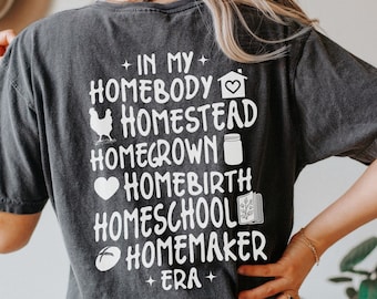 May include: A dark gray t-shirt with white text that reads "In My Homebody Homestead Homegrown Homebirth Homeschool Homemaker Era".