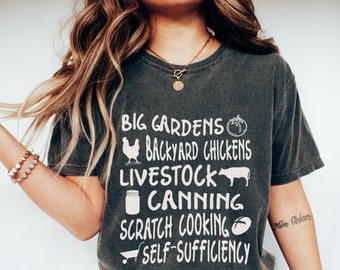 May include: A dark gray t-shirt with white text that reads "Big Gardens, Backyard Chickens, Livestock, Canning, Scratch Cooking, Self-Sufficiency". The text is accompanied by small icons of a tomato, chicken, cow, jar, rolling pin, and a shopping cart.