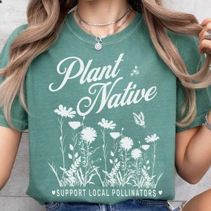 May include: A teal green t-shirt with a white graphic that says "Plant Native" with a butterfly and bee design. The graphic also includes a line of white flowers and the text "Support Local Pollinators".
