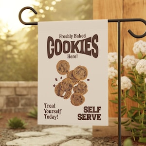 May include: A white garden flag with brown text that reads "Freshly Baked Cookies Here!" and "Treat Yourself Today!"  The flag also features a graphic of chocolate chip cookies and the text "Self Serve".