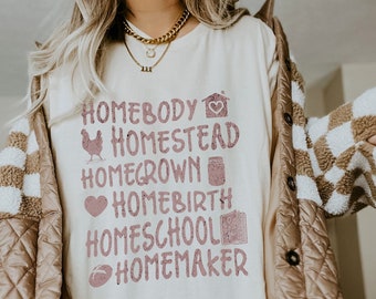 May include: A white t-shirt with a pink graphic that says "Homebody Homestead Homegrown Homebirth Homeschool Homemaker". The graphic includes icons for a house, a chicken, a jar, a heart, a book, and a football.