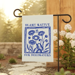 May include: A cream-colored garden flag with a blue floral design and the text "PLANT NATIVE FOR POLLINATORS." The flag is hanging on a black metal stand in a garden setting. The design features stylized flowers and a butterfly.
