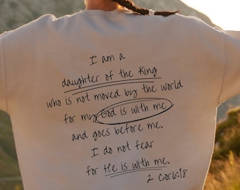 May include: A person wearing a white sweatshirt with the text "I am a daughter of the King who is not moved by the world for my God is with me and goes before me. I do not fear for He is with me. 2 Cor 6:18" written on the back. The person is standing with their arms raised in the air, looking out at a mountain range in the distance.