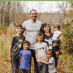 May include: A family of six, including two parents and four children, stand in a field of tall grass. They are all wearing t-shirts with various designs related to farm life. The shirts feature phrases like "Life is better on the homestead" and "Chicken Whisperer".