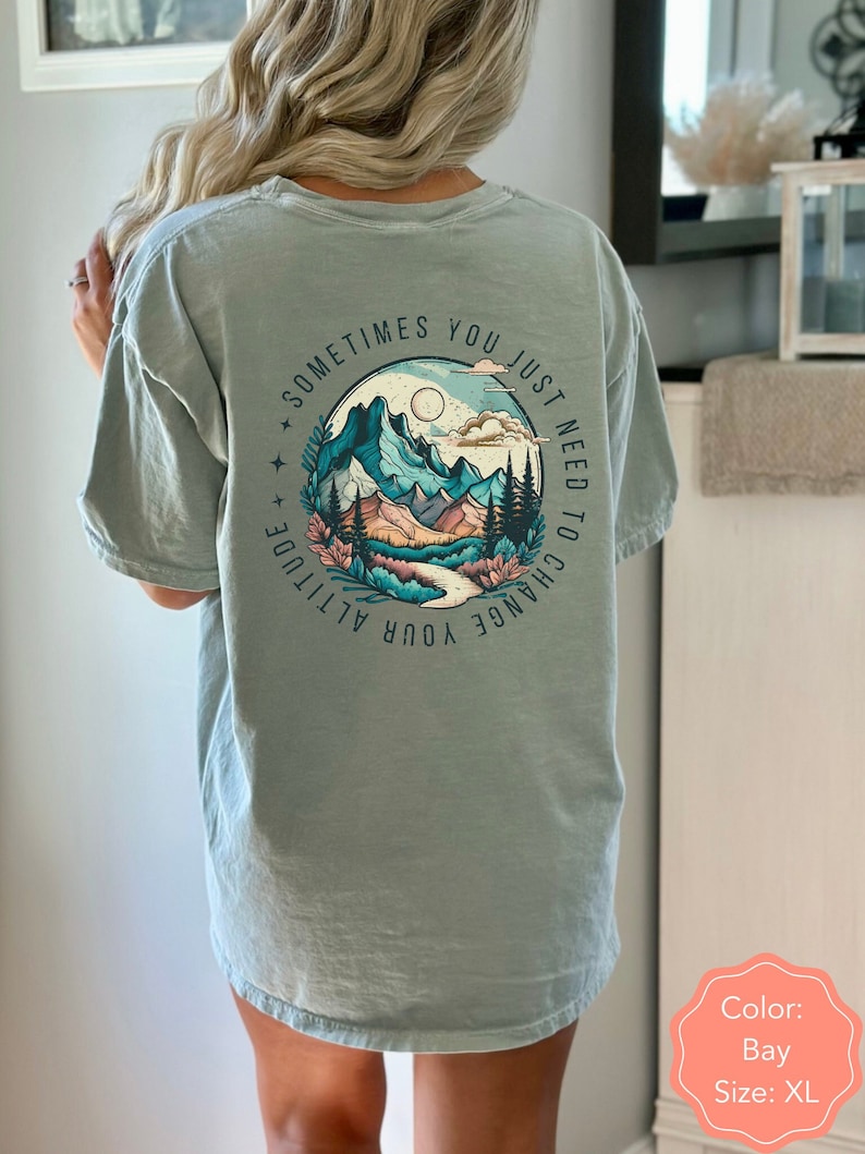 May include: A light blue t-shirt with a graphic of mountains, a river, and trees. The text "Sometimes you just need to change your attitude" is printed around the graphic.