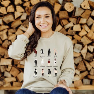 K&ouml;nnte beinhalten: Eine Frau tr&auml;gt einen hellbeigen Pullover mit einer Grafik verschiedener Pferderassen. Der Pullover hat die Namen der einzelnen Rassen unter dem Bild aufgedruckt. Die Frau steht vor einer Wand aus gestapeltem Brennholz.