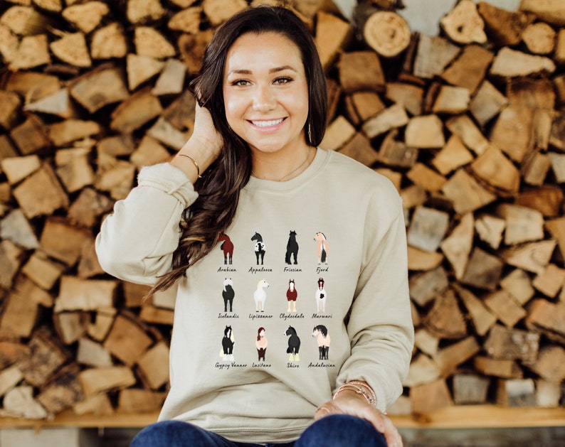 K&ouml;nnte beinhalten: Eine Frau tr&auml;gt einen hellbeigen Pullover mit einer Grafik verschiedener Pferderassen. Der Pullover hat die Namen der einzelnen Rassen unter dem Bild aufgedruckt. Die Frau steht vor einer Wand aus gestapeltem Brennholz.