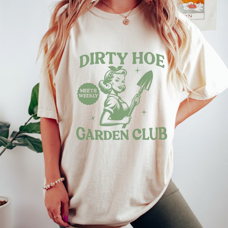 May include: A white t-shirt with a green graphic design that says "Dirty Hoe Meets Weekly Garden Club". The graphic features a woman holding a shovel.