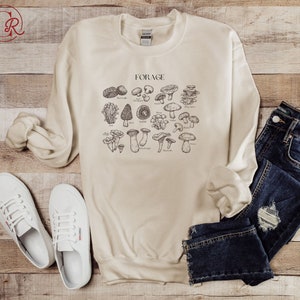 May include: A beige sweatshirt with a graphic of various types of mushrooms and the word "FORAGE" printed on the front. The sweatshirt is laid out on a wooden surface with a pair of white sneakers and a pair of blue jeans.