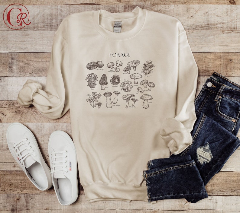 May include: A beige sweatshirt with a graphic of various types of mushrooms and the word "FORAGE" printed on the front. The sweatshirt is laid out on a wooden surface with a pair of white sneakers and a pair of blue jeans.