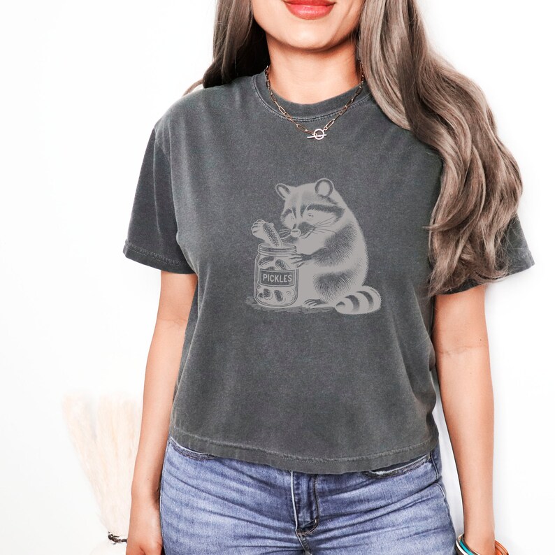 May include: A dark gray cropped t-shirt with a graphic of a raccoon holding a jar of pickles. The jar says "Pickles" on the label.