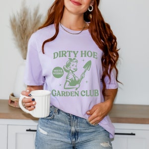 May include: A purple t-shirt with the text "Dirty Hoe Meets Weekly Garden Club" and a green illustration of a woman holding a shovel.