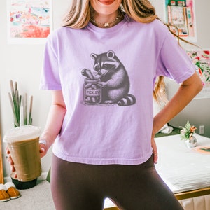 May include: A purple t-shirt with a graphic of a raccoon holding a jar of pickles. The jar says "Pickles" on it.