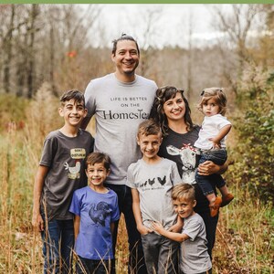 Pode incluir: Uma fam&iacute;lia de seis pessoas, incluindo dois pais e quatro filhos, est&aacute; em p&eacute; em um campo de grama alta. Todos est&atilde;o vestindo camisetas com v&aacute;rios designs relacionados &agrave; vida na fazenda. As camisetas apresentam frases como "A vida &eacute; melhor na fazenda" e "Sussurrador de galinhas".