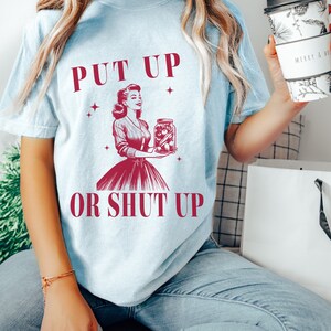 May include: Light blue t-shirt with the text "PUT UP OR SHUT UP" and a vintage illustration of a woman holding a jar of preserved food. The shirt is a light blue color.