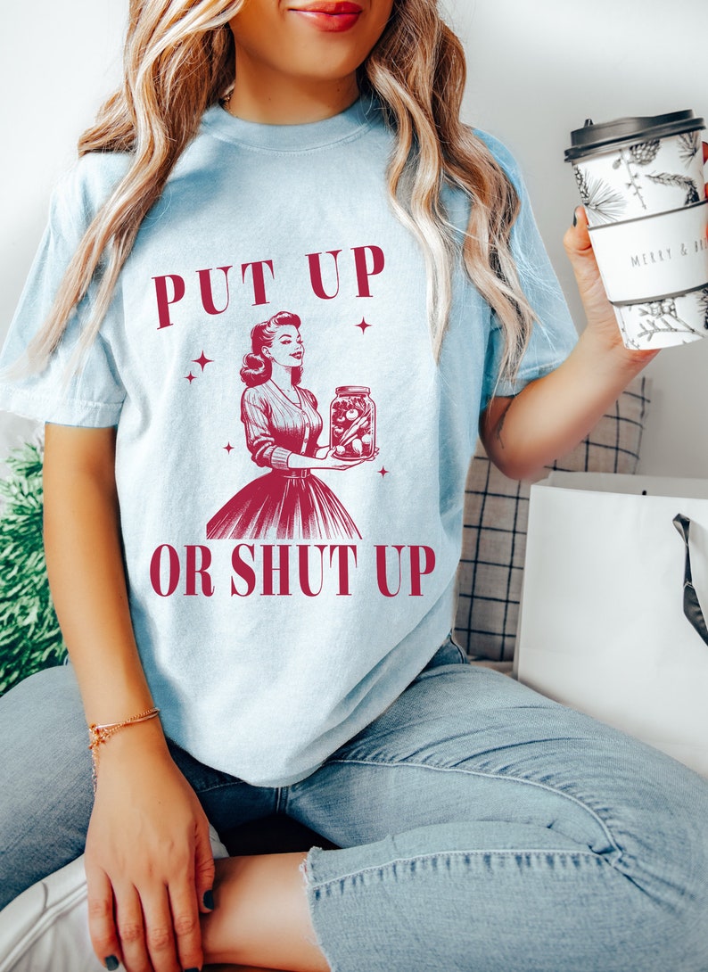 May include: Light blue t-shirt with the text "PUT UP OR SHUT UP" and a vintage illustration of a woman holding a jar of preserved food. The shirt is a light blue color.