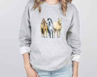 May include: A gray sweatshirt with a watercolor illustration of three horses. The horses are standing side-by-side, with the gray horse in the middle and the brown and white horses on either side.