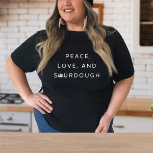 Pode incluir: Uma mulher vestindo uma camiseta preta com o texto "Peace, Love, and Sourdough" em letras brancas. Ela est&aacute; em p&eacute; em uma cozinha com uma bancada e arm&aacute;rios no fundo.