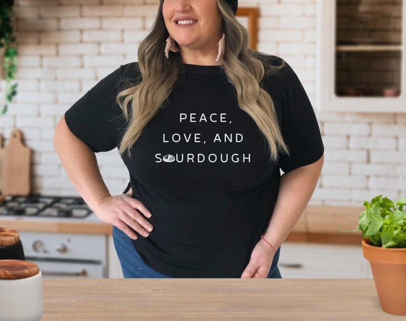 Pode incluir: Uma mulher vestindo uma camiseta preta com o texto "Peace, Love, and Sourdough" em letras brancas. Ela est&aacute; em p&eacute; em uma cozinha com uma bancada e arm&aacute;rios no fundo.