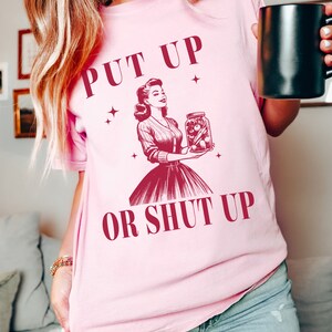 May include: Pink t-shirt with the text "PUT UP OR SHUT UP" and a vintage illustration of a woman holding a jar of pickles. The design is in a deep red color. The shirt is a light pink color.