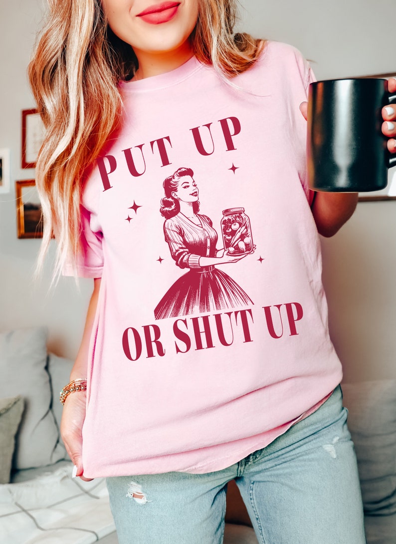 May include: Pink t-shirt with the text "PUT UP OR SHUT UP" and a vintage illustration of a woman holding a jar of pickles. The design is in a deep red color. The shirt is a light pink color.