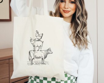 May include: A white canvas tote bag with a black and white illustration of a cow, pig, and chicken stacked on top of each other. The illustration is in a vintage style.