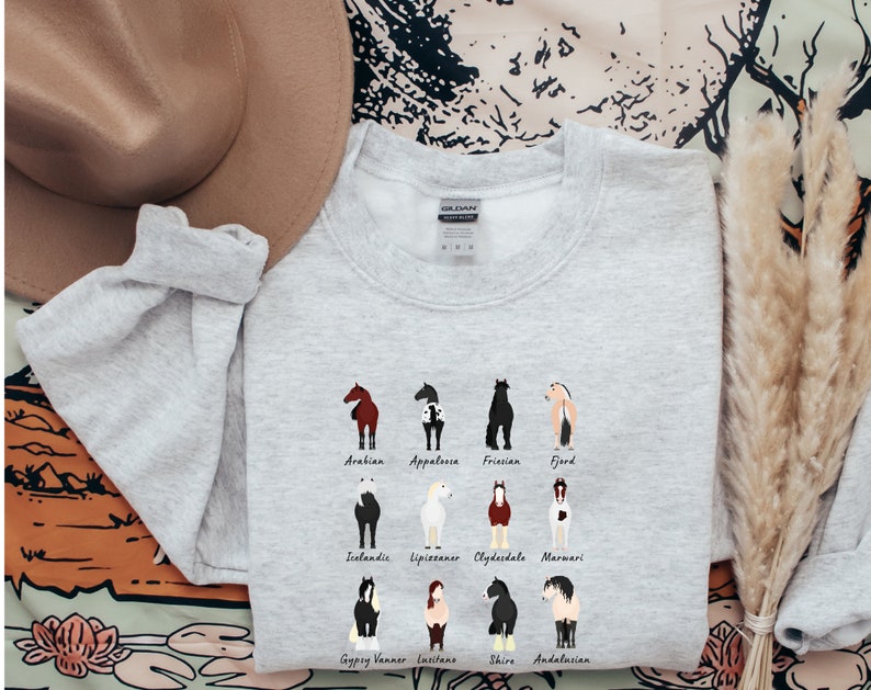K&ouml;nnte beinhalten: Grauer Pullover mit einer Grafik verschiedener Pferderassen, darunter Araber, Appaloosa, Friese, Fjord, Isl&auml;nder, Lipizzaner, Clydesdale, Marwari, Gypsy Vanner, Lusitano, Shire und Andalusier.