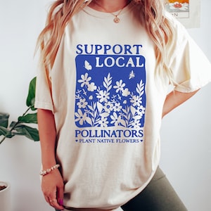 May include: A cream-colored t-shirt with a blue graphic that says "Support Local Pollinators Plant Native Flowers". The graphic features a stylized floral design with butterflies and bees.