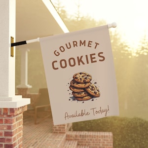 May include: A tan fabric flag with the text "Gourmet Cookies" in brown lettering. A stack of chocolate chip cookies is pictured below the text. The text "Available Today!" is at the bottom of the flag.