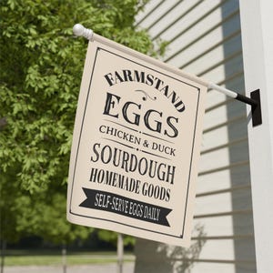 Peut inclure: Un drapeau rectangulaire crème avec du texte noir indiquant "FARMSTAND EGGS CHICKEN & DUCK SOURDOUGH HOMEMADE GOODS SELF-SERVE EGGS DAILY." Le drapeau est suspendu à l'extérieur sur un bâtiment blanc.