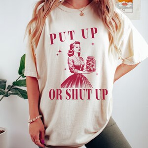 May include: Cream-colored t-shirt with the text "PUT UP OR SHUT UP" in burgundy. The graphic features a vintage illustration of a woman holding a jar of pickles. The shirt has a relaxed fit.