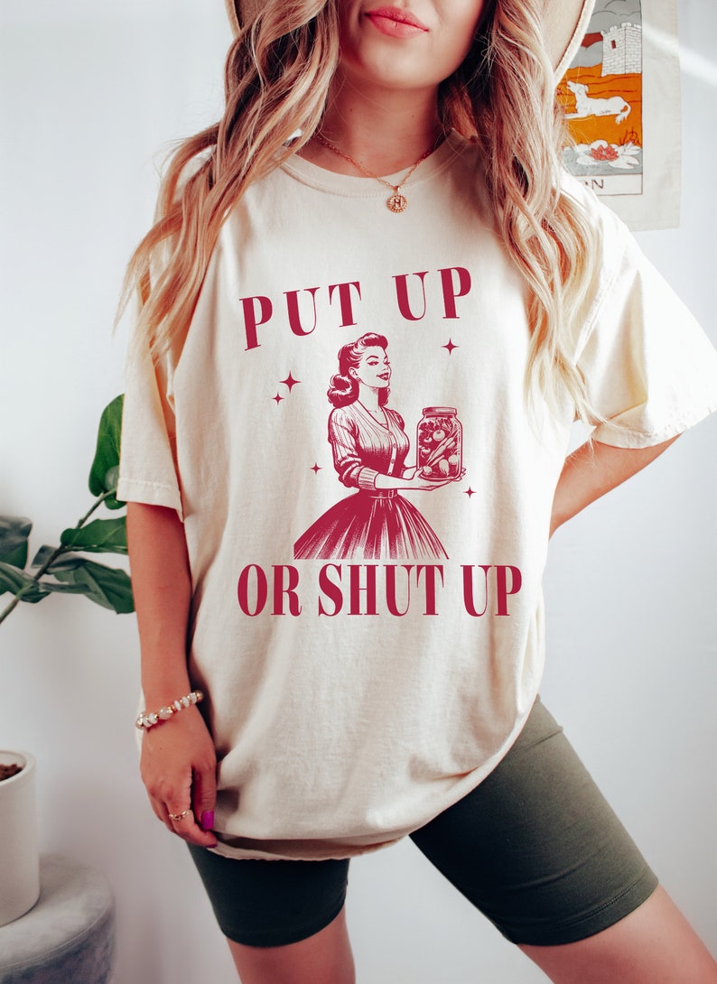 May include: Cream-colored t-shirt with the text "PUT UP OR SHUT UP" in burgundy. The graphic features a vintage illustration of a woman holding a jar of pickles. The shirt has a relaxed fit.