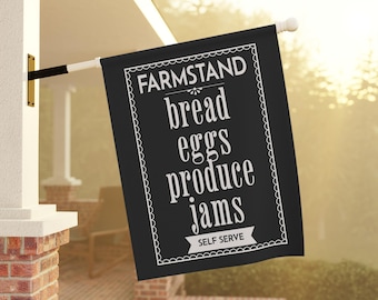 May include: A black rectangular flag with white text that reads "FARMSTAND bread eggs produce jams SELF SERVE". The flag is hanging outside a building.