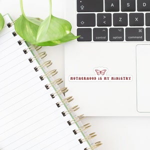 May include: A white laptop with a sticker that says "Motherhood is my ministry" with a red butterfly design. The laptop is on a white surface with a notebook and a pen.