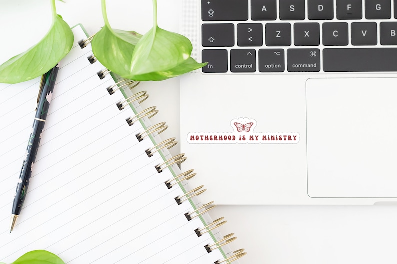 May include: A white laptop with a sticker that says "Motherhood is my ministry" with a red butterfly design. The laptop is on a white surface with a notebook and a pen.
