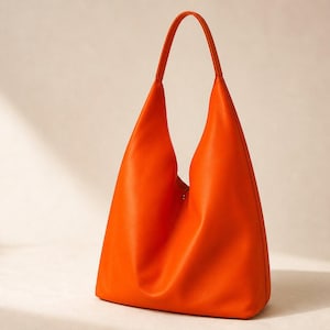 May include: A vibrant orange hobo bag featuring a smooth, solid-coloured exterior, a single integrated shoulder strap, and a deep, slouchy silhouette with a central magnetic snap closure visible at the top opening of the main compartment.