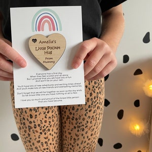 Pu&ograve; includere: Un'etichetta di legno a forma di cuore con il testo "Amelia's Little Pocket Hug From Mummy". L'etichetta &egrave; attaccata a una carta con un design arcobaleno e un messaggio sul primo giorno di un bambino.