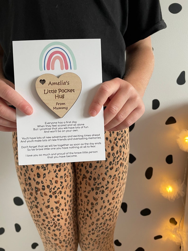 Pu&ograve; includere: Un'etichetta di legno a forma di cuore con il testo "Amelia's Little Pocket Hug From Mummy". L'etichetta &egrave; attaccata a una carta con un design arcobaleno e un messaggio sul primo giorno di un bambino.