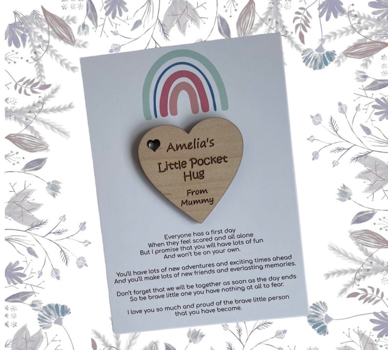 Pu&ograve; includere: Un portafortuna di legno a forma di cuore con il testo "Amelia's Little Pocket Hug From Mummy" su una carta bianca con un'illustrazione di arcobaleno. La carta include una poesia sul primo giorno di un bambino.