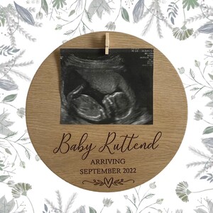 May include: A round wooden sign with a photo of a baby's ultrasound scan. The sign is decorated with a floral design and the text "Baby Ruttend Arriving September 2022".