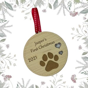 May include: A round wooden ornament with the text "Jasper's First Christmas 2021" and a paw print. Two small silver hearts are above the paw print. The ornament is attached to a red ribbon with white polka dots.