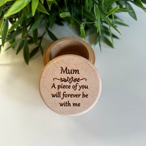 Può includere: Piccola scatola di legno aperta con coperchio. Il coperchio è inciso con la parola "Mum" sopra la frase "A piece of you will forever be with me". Sullo sfondo è visibile del fogliame verde.