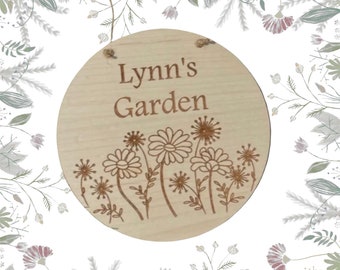 Könnte beinhalten: Ein rundes Holzschild mit dem Text "Lynn's Garden" und einem Design aus stilisierten Blumen. Das Schild hat ein Seil zum Aufhängen.