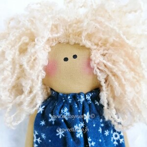 Peut inclure: Une poupée faite à la main avec des cheveux blonds bouclés portant une robe bleue avec des flocons de neige blancs. La poupée a un visage beige avec des joues roses et des yeux noirs. La robe porte le texte "www.mittsyrums.com @sweettalojh".